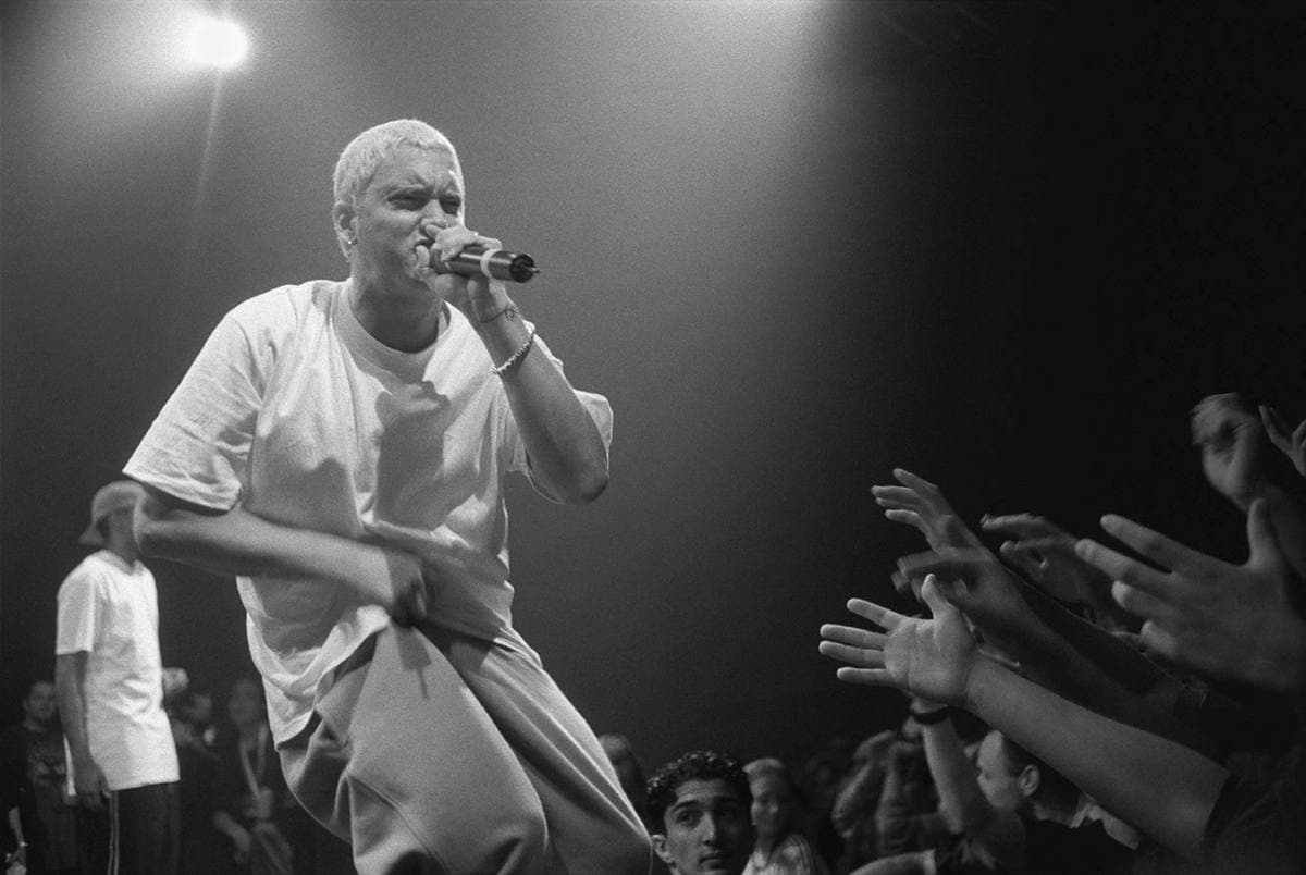 Eminem in a white T-shirt crouching at the edge of a stage with a microphone as fans reach towards him