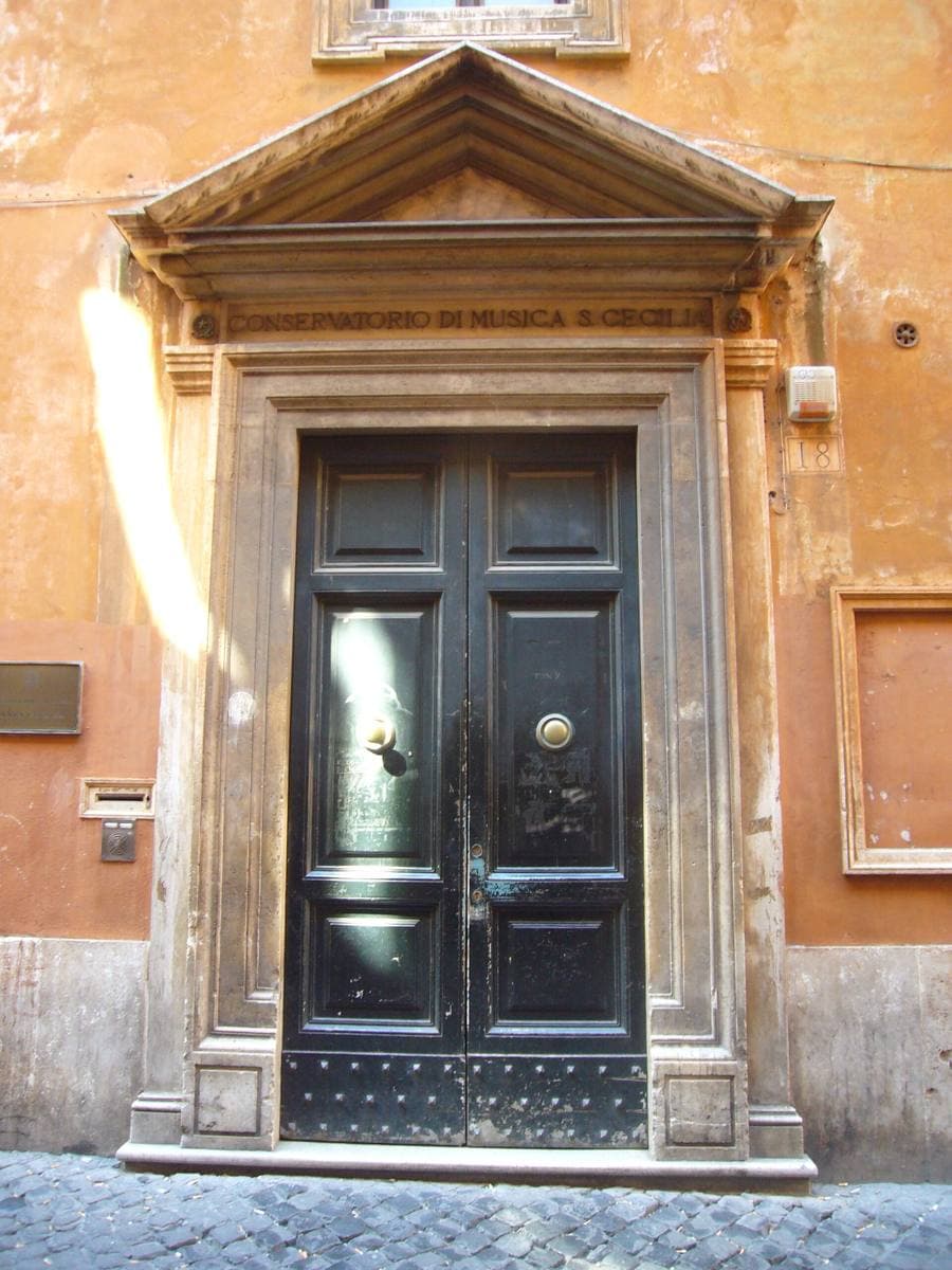 A tall black door under a stone pediment carved Conservatorio di Musica S. Cecilia, in an ochre wall