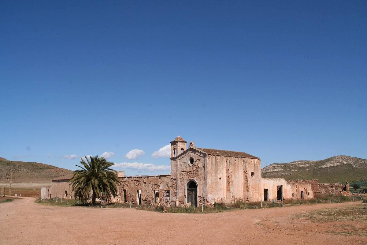 A ruined pink farmhouse and chapel with a palm tree, under a deep blue sky