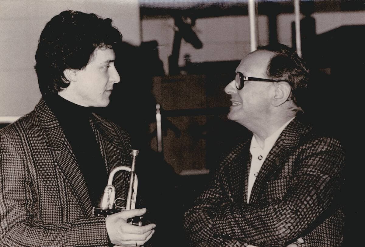 Ennio Morricone in glasses and a checked jacket, smiling at a younger trumpeter holding his instrument