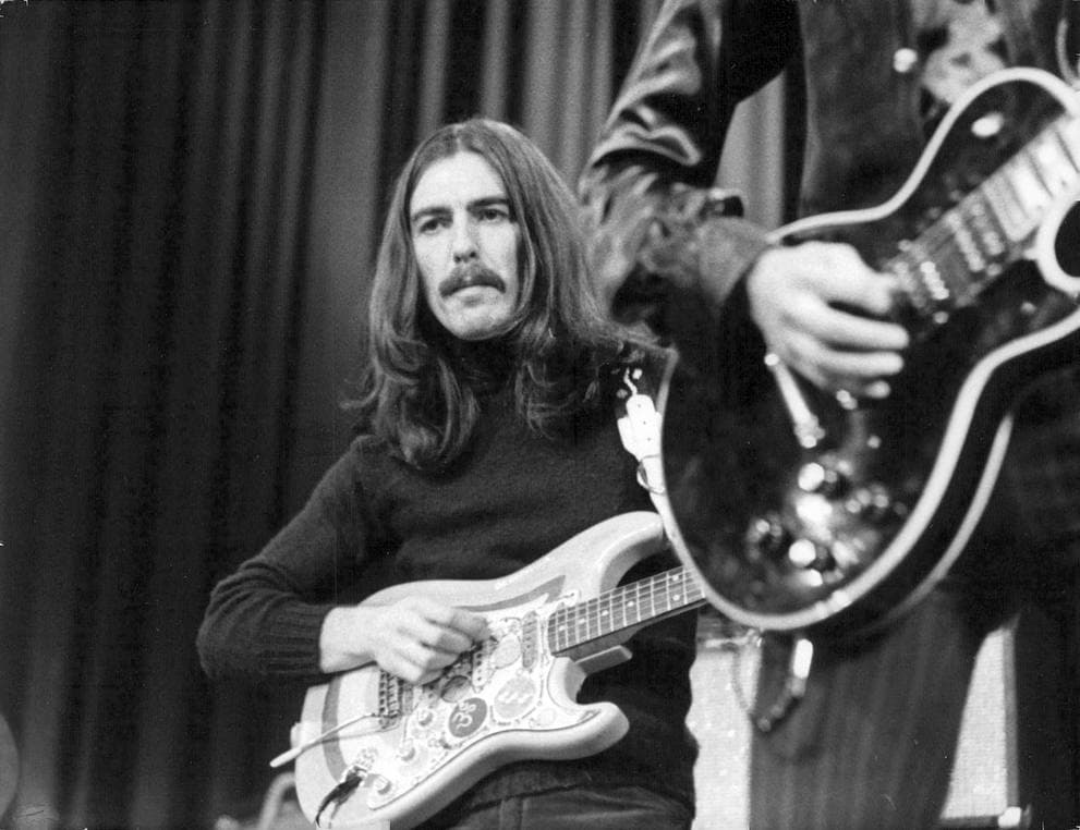 George Harrison in a dark jumper playing a painted electric guitar, another guitarist's hands at the edge of the frame