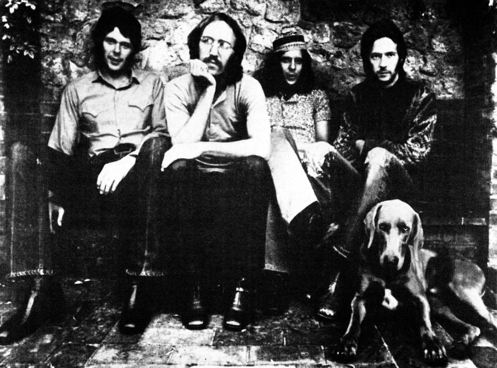 Four bearded men sitting against a stone wall with a dog beside them