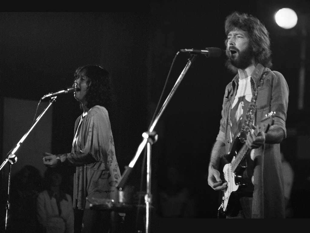 Yvonne Elliman and Eric Clapton singing at two microphones on a dark stage
