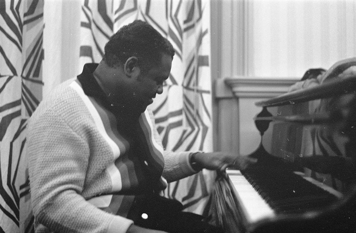 Fats Domino in a striped cardigan playing an upright piano in a hotel room