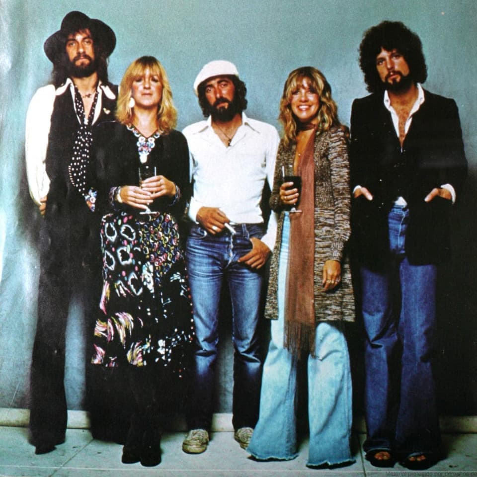 The five members of Fleetwood Mac standing in a row against a pale blue wall, Stevie Nicks in a long fringed scarf