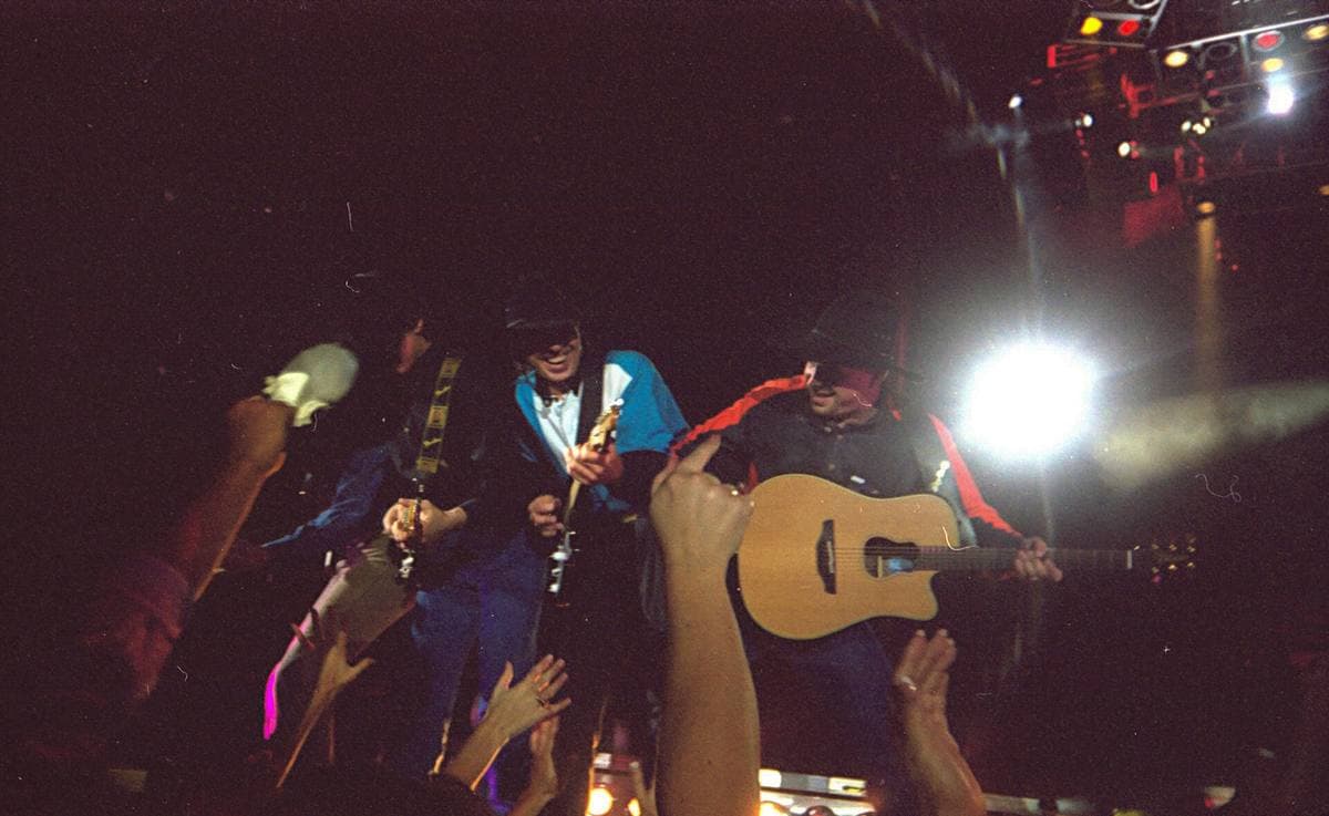 Garth Brooks playing an acoustic guitar under stage lights, fans’ hands reaching up in front of him