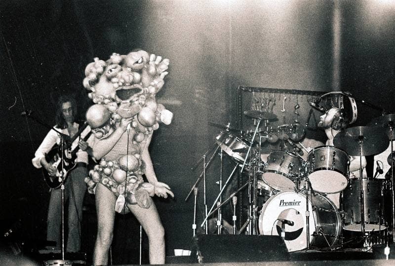 A figure in a lumpy full-body costume on a stage beside a drum kit and guitarist