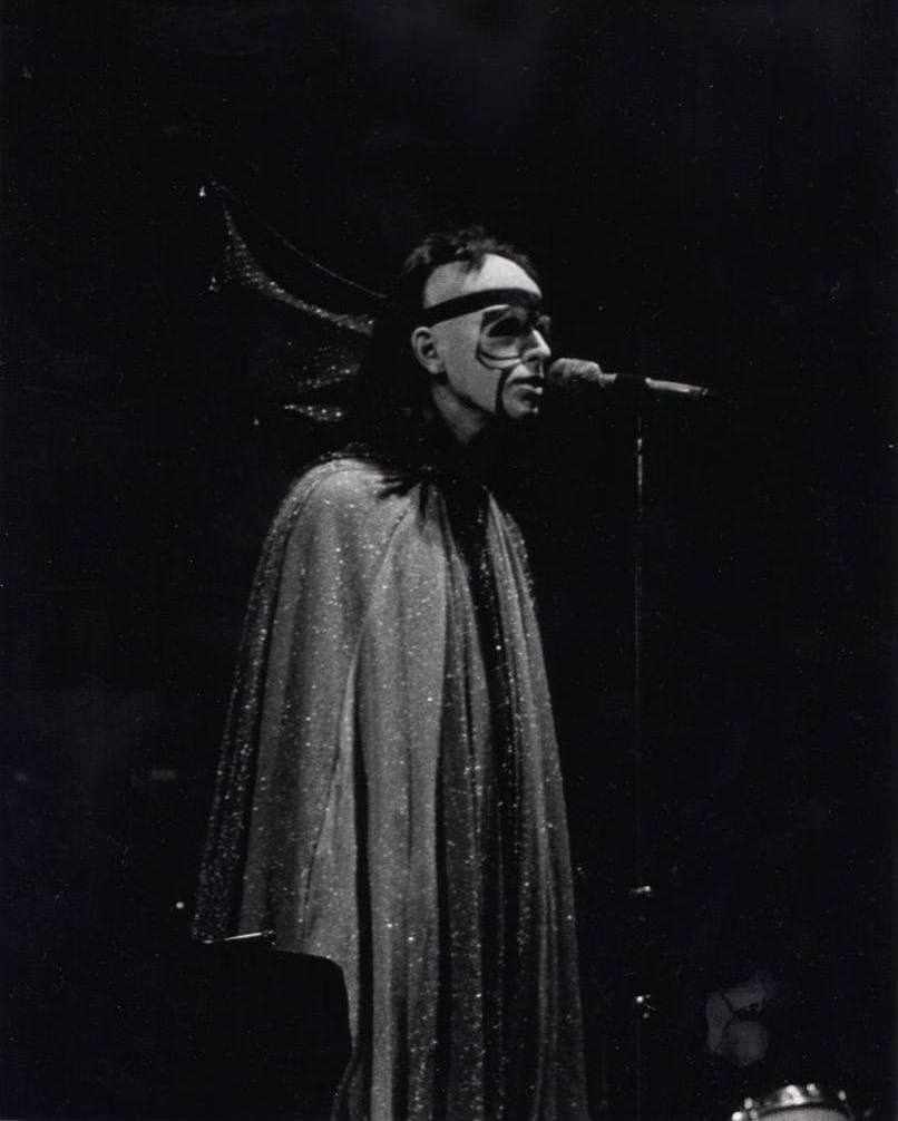 A singer in a glittering cape, white face paint and a bat-wing headdress, at a microphone on a dark stage