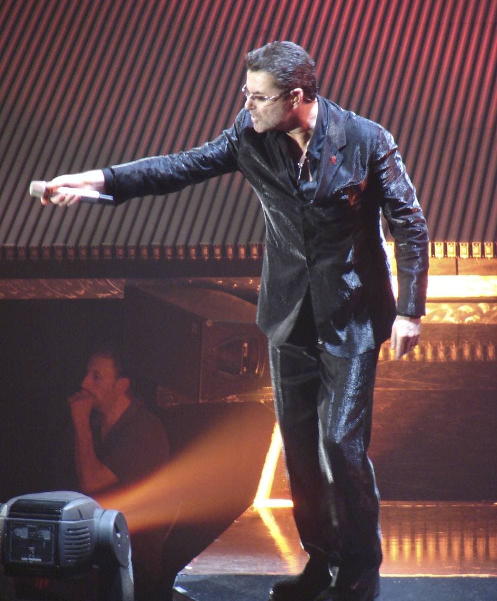 George Michael on stage in a glossy black suit and tinted glasses, reaching out with the microphone towards the crowd