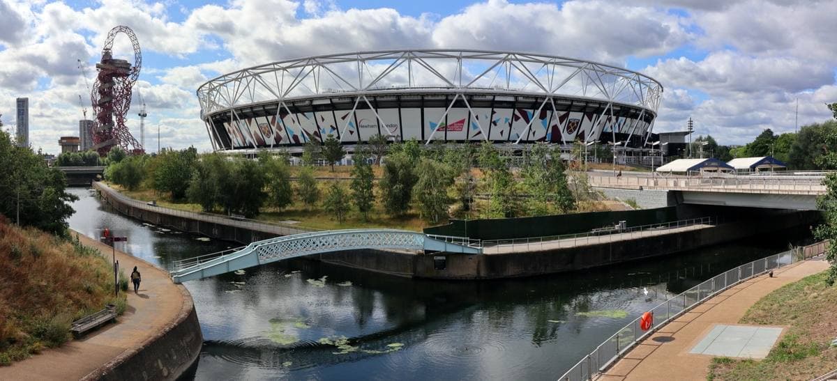 The bowl of London Stadium beyond a river and footbridge in the Olympic Park, the red Orbit tower to one side
