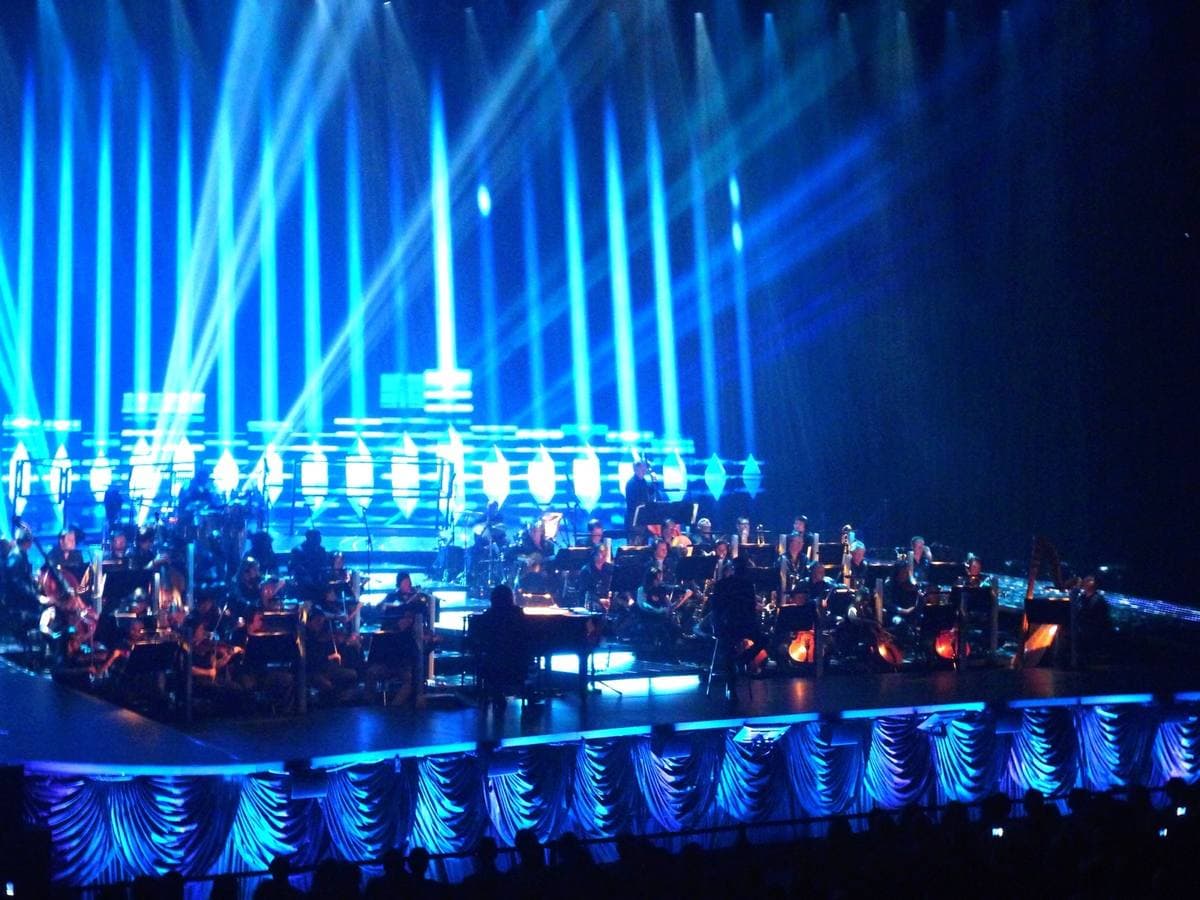 An orchestra on a tiered stage under blue light beams, a grand piano at the front