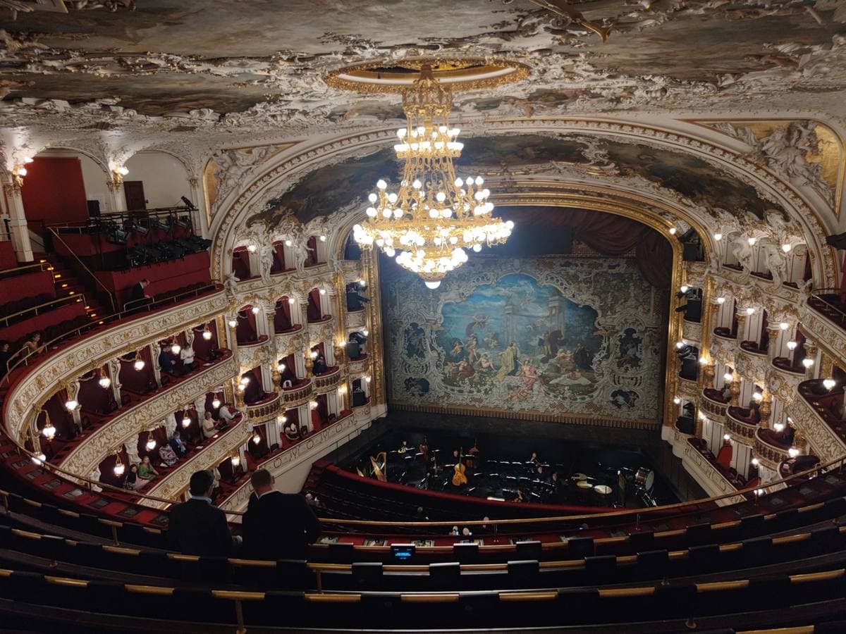 A red and gold horseshoe auditorium with tiers of boxes, a crystal chandelier and a painted curtain