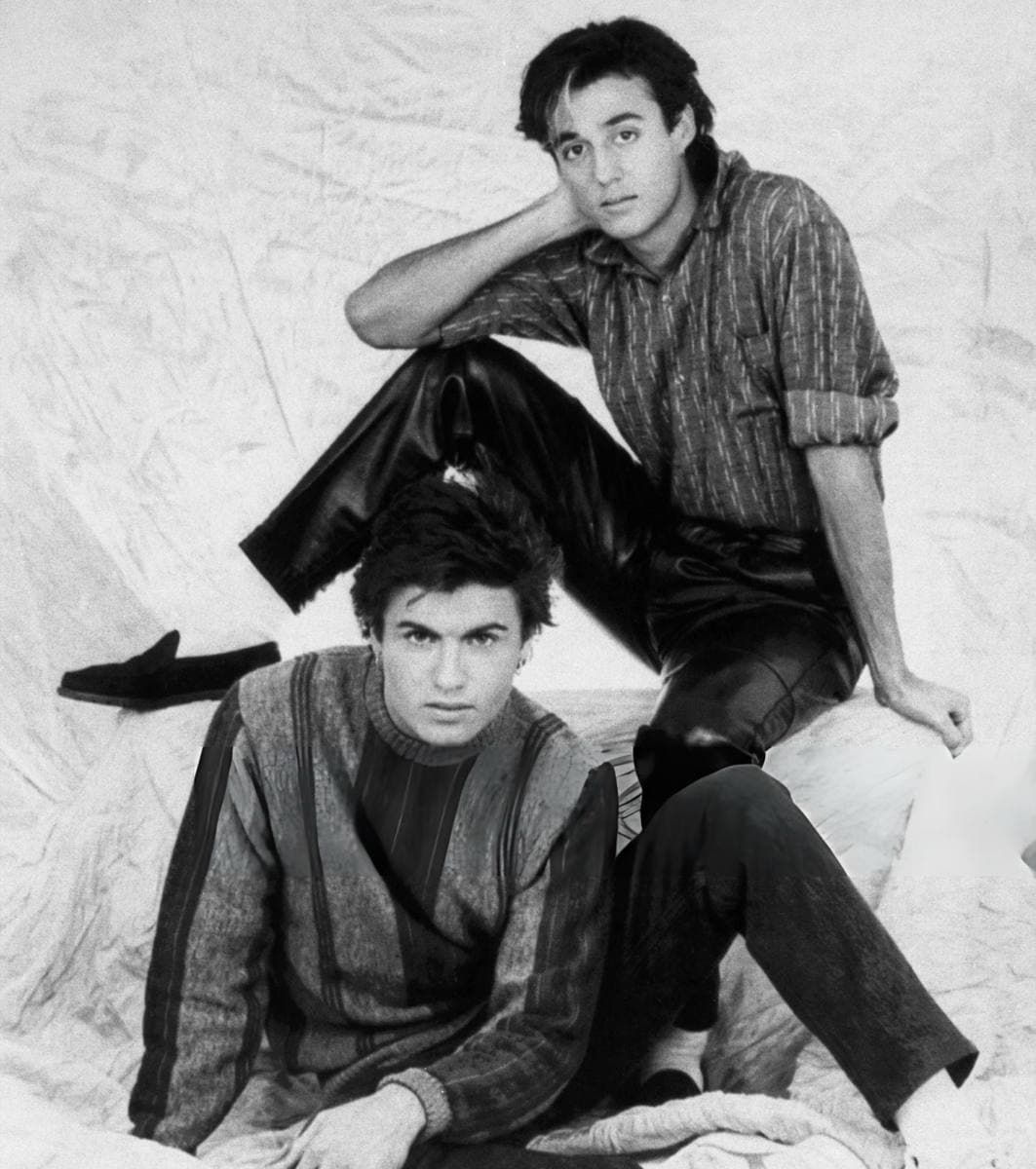 Two young men posed on a pale draped backdrop, Andrew Ridgeley seated behind in a checked shirt, George Michael in front in a patterned jumper