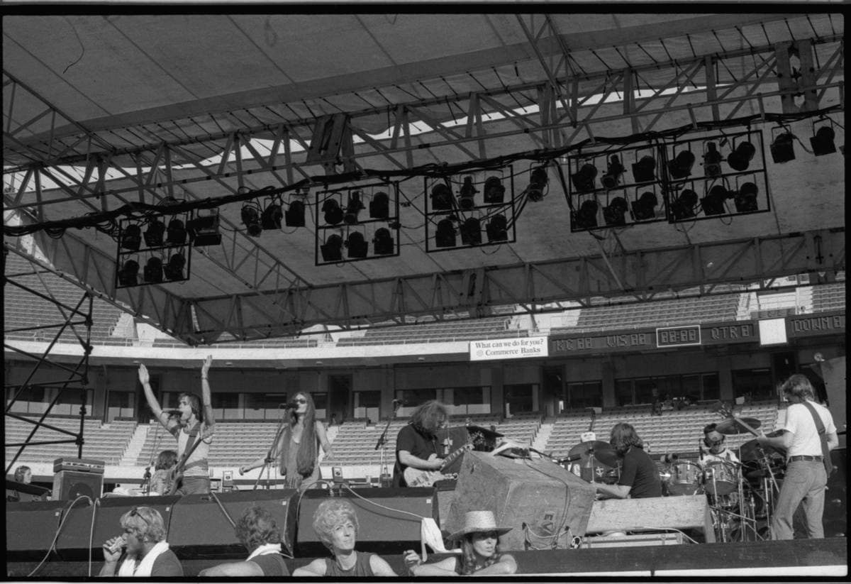 A black-and-white view of the band on a big open-air stadium stage under a lighting rig, fans in front