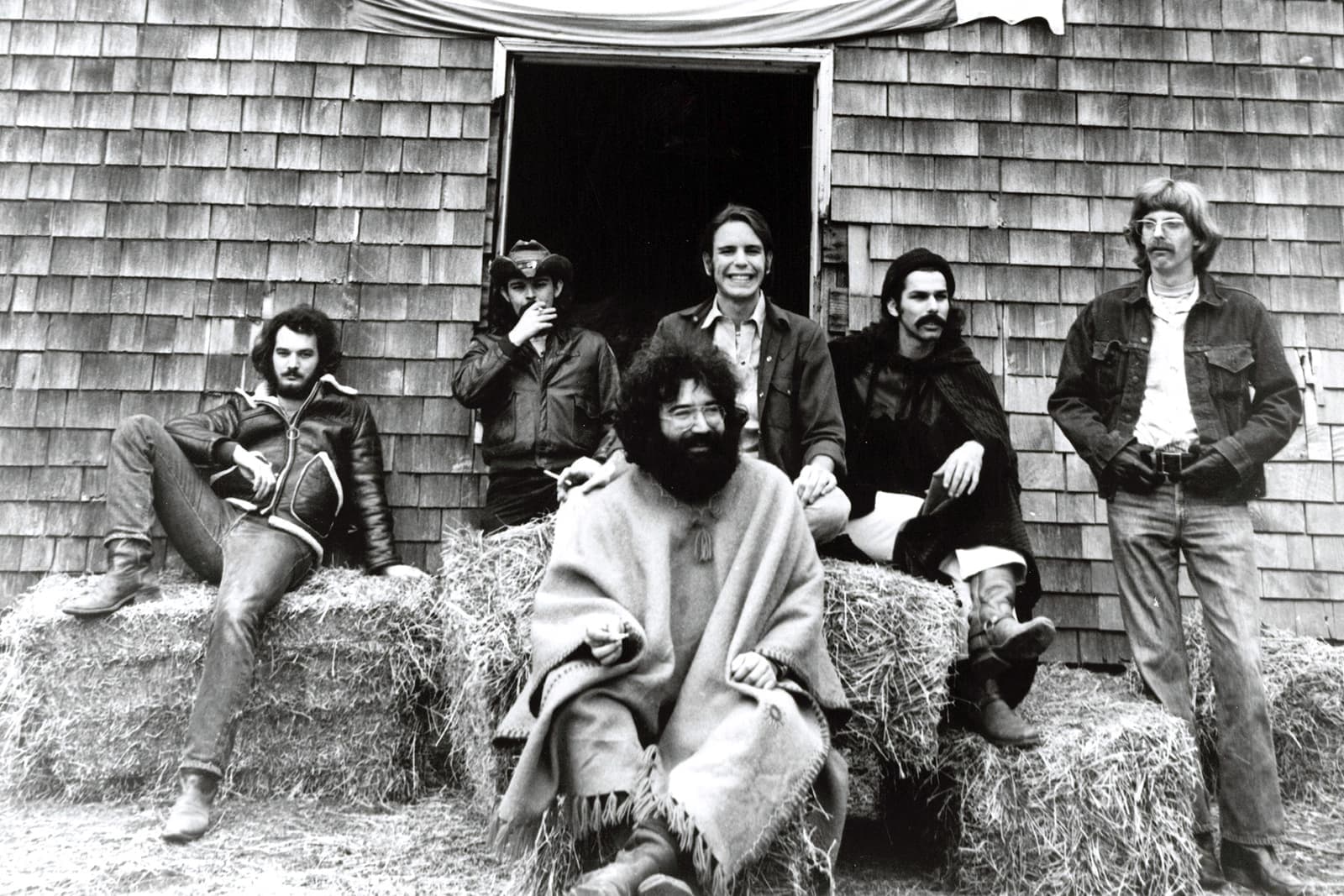 Six men in front of a shingled barn: Garcia seated centre in a poncho, the others on hay bales and in the doorway