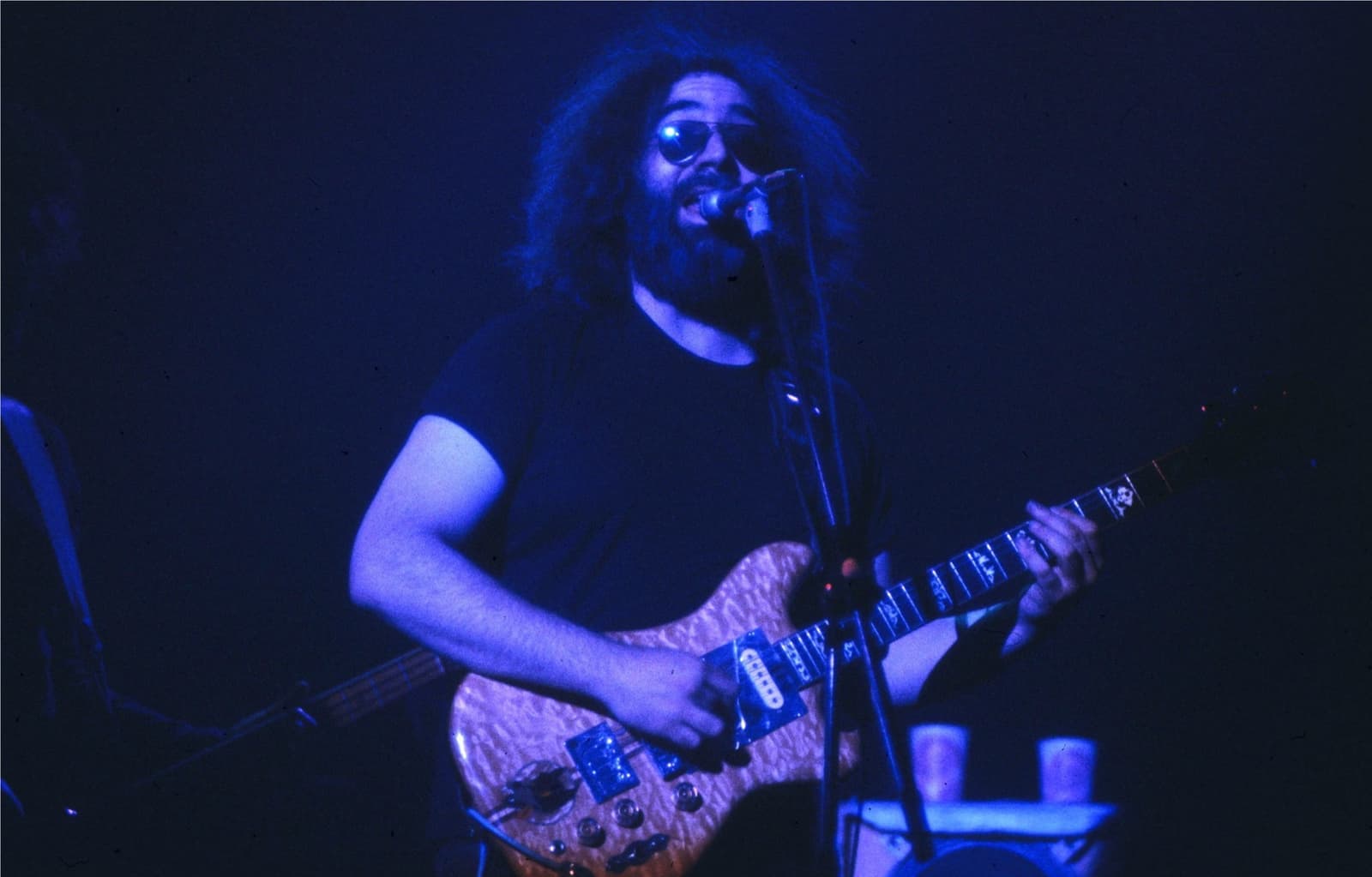 Jerry Garcia in dark glasses singing at a microphone under blue stage light, a wooden guitar in his hands