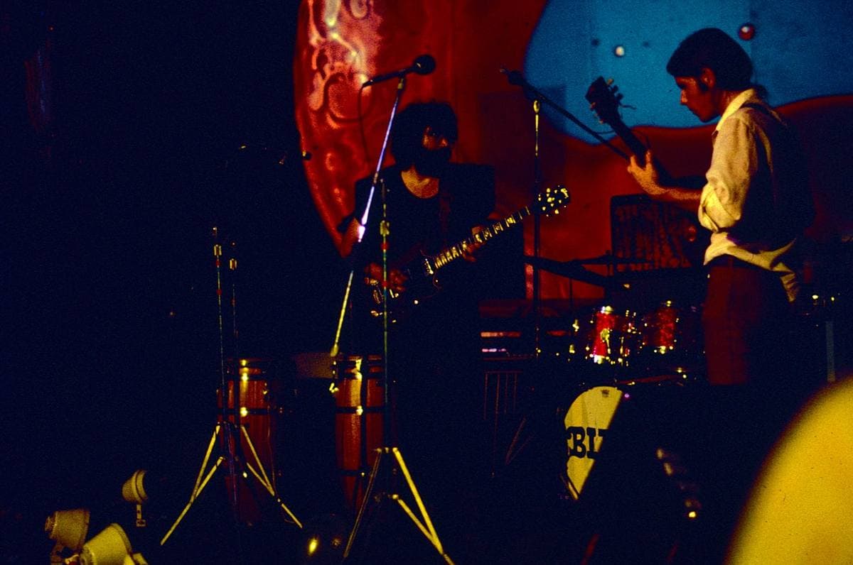 The band on a dark stage in red and blue light, a huge projected liquid blob glowing behind them