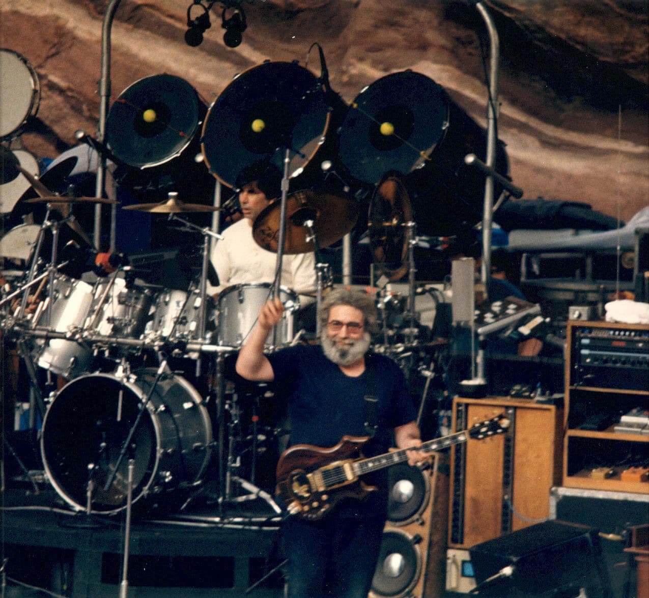 Jerry Garcia raising a hand in front of Mickey Hart’s drum kit, red sandstone behind