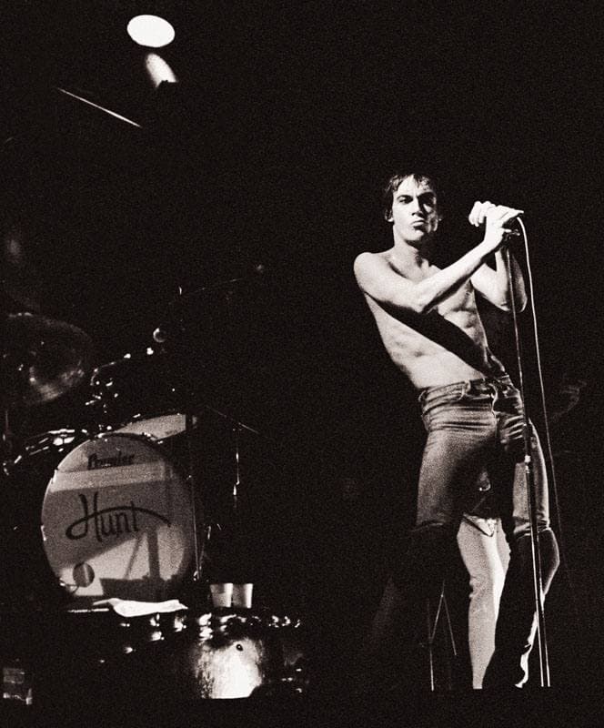 Iggy Pop shirtless in black and white, gripping the microphone stand under a single spotlight