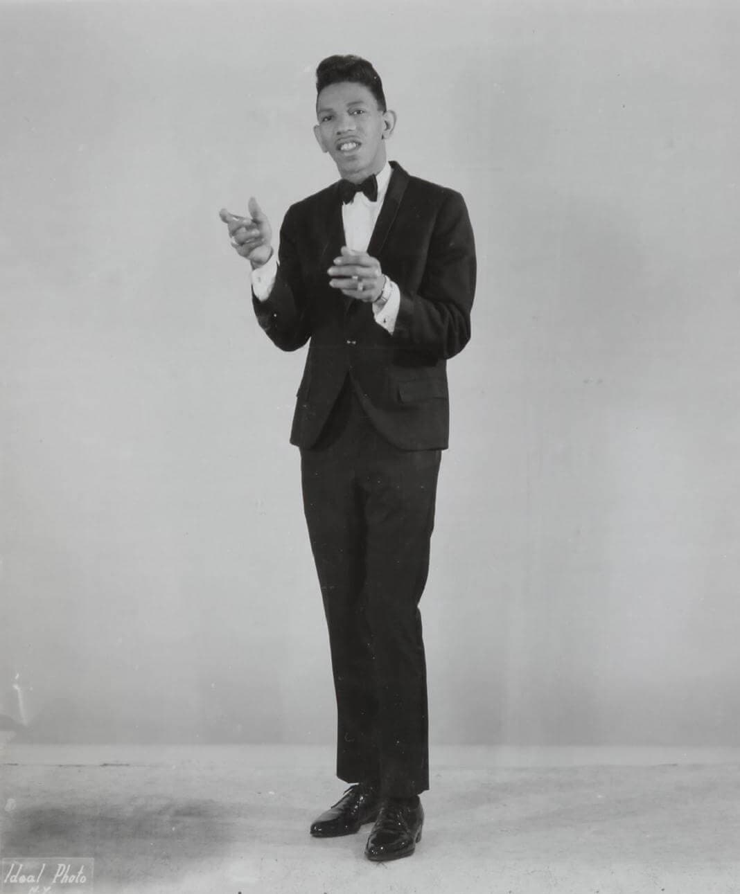Bobby Byrd in a dark tuxedo and bow tie, standing and gesturing with both hands
