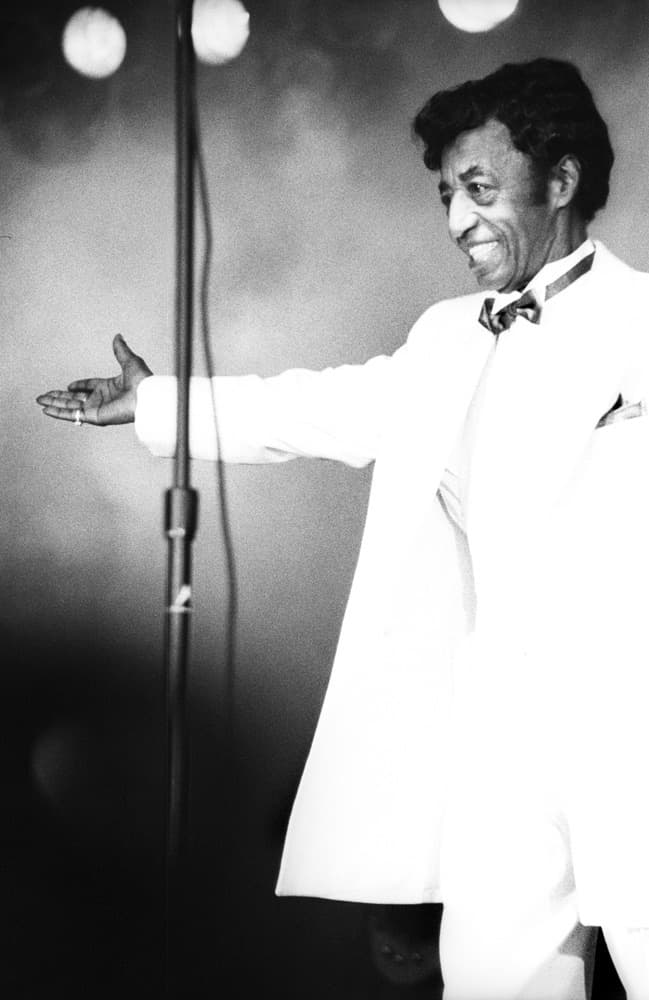 Danny Ray in a white tuxedo, arm out towards the stage, in a spotlight