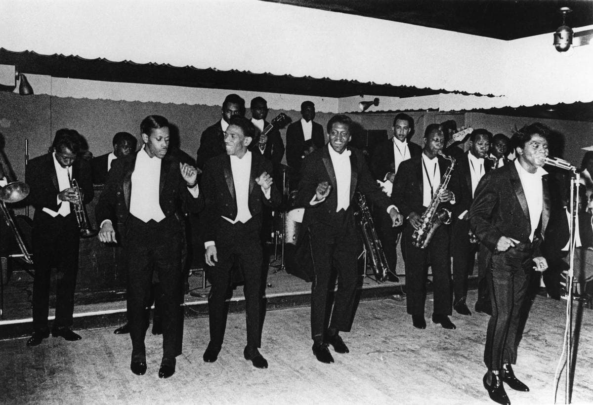 James Brown at the microphone in a dark suit, a line of dancing singers and horn players in white shirts behind him