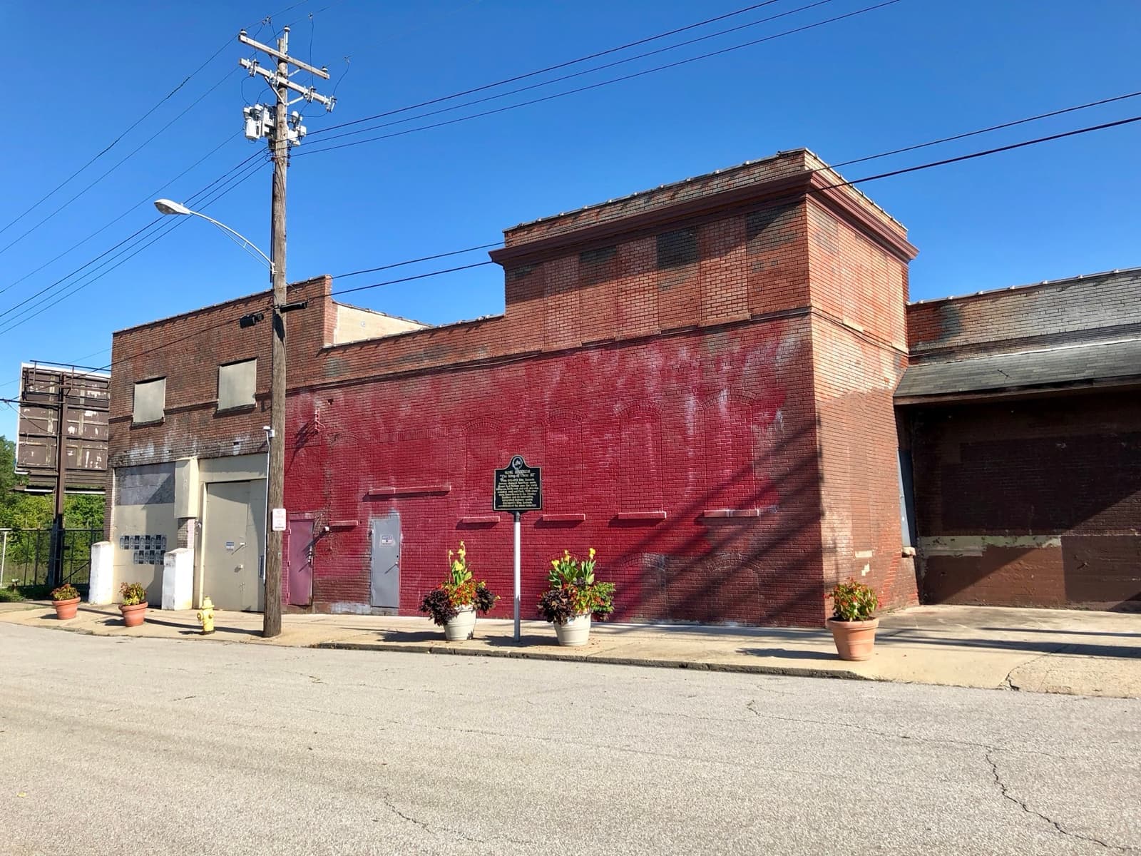 A red-brick industrial building with a historical marker and plant pots in front