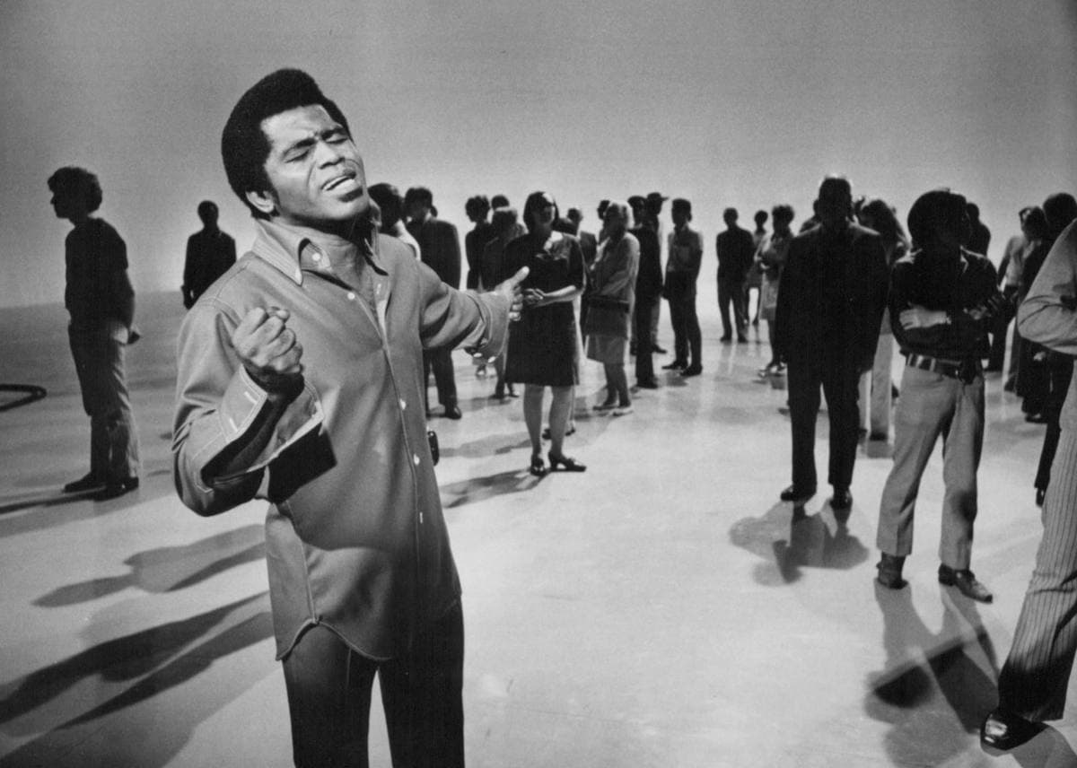 James Brown singing with eyes closed and fist clenched on a white studio floor, dancers behind him