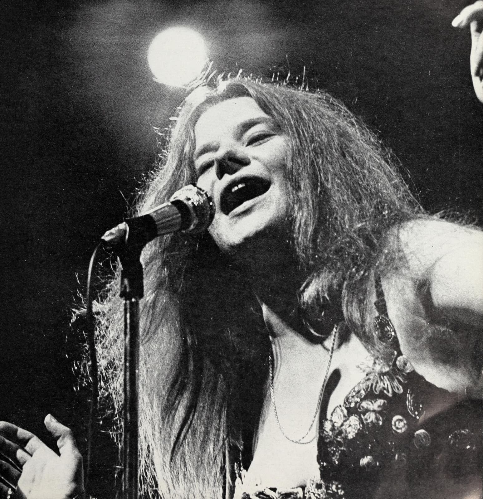 Janis Joplin singing into a microphone under a spotlight, head tilted back, long hair loose