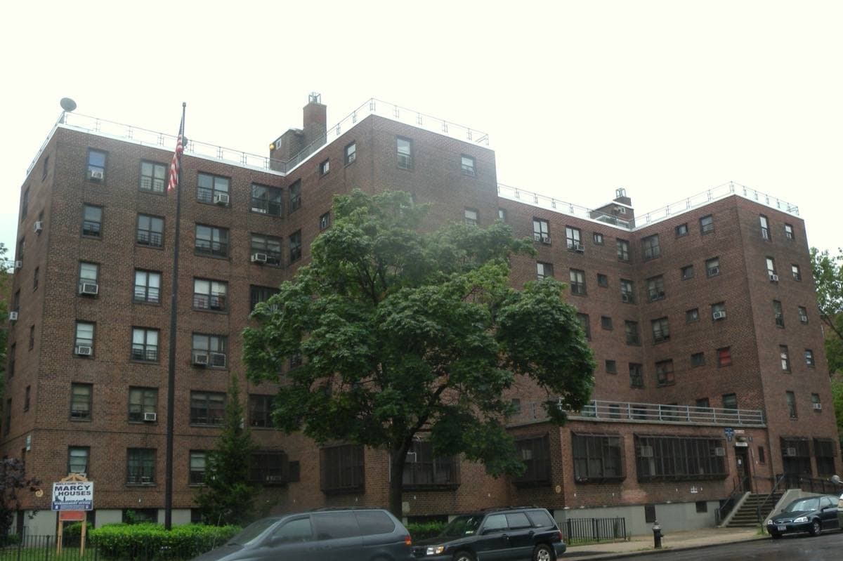 Six-storey brick buildings of Marcy Houses behind trees on a wet street