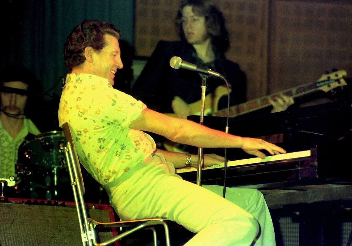 Jerry Lee Lewis in a bright shirt playing a piano on stage, a bassist behind him
