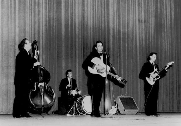 Johnny Cash with guitar, a bass player, a drummer and an electric guitarist on a curtained stage