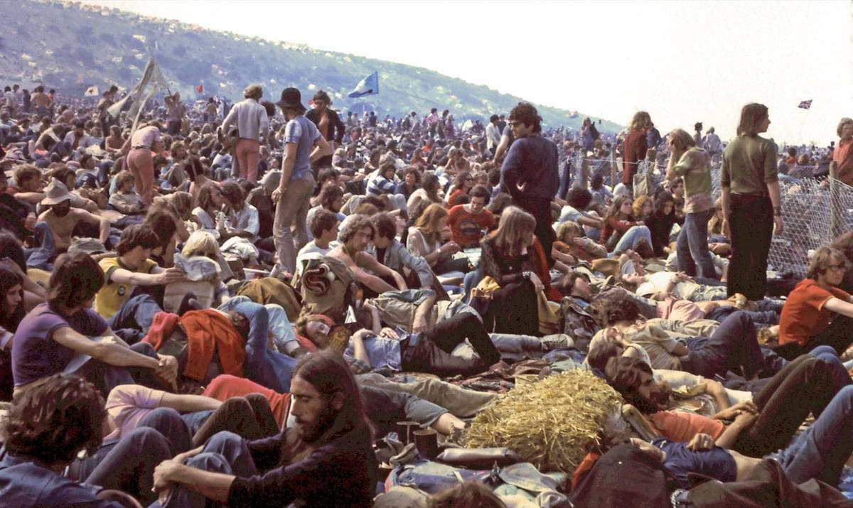 A dense crowd sitting and standing on a grassy slope under a hill