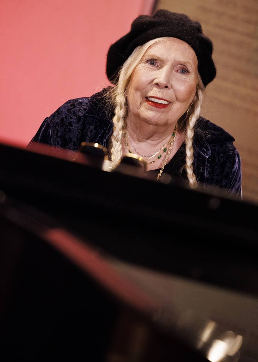 Joni Mitchell in a beret and dark glasses seated at a grand piano