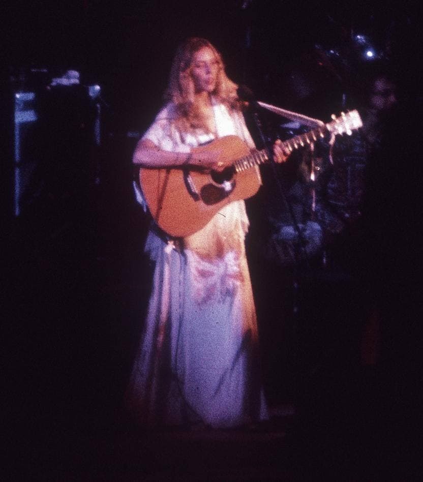 Joni Mitchell on a dark stage in a long pale dress, playing an acoustic guitar at a microphone