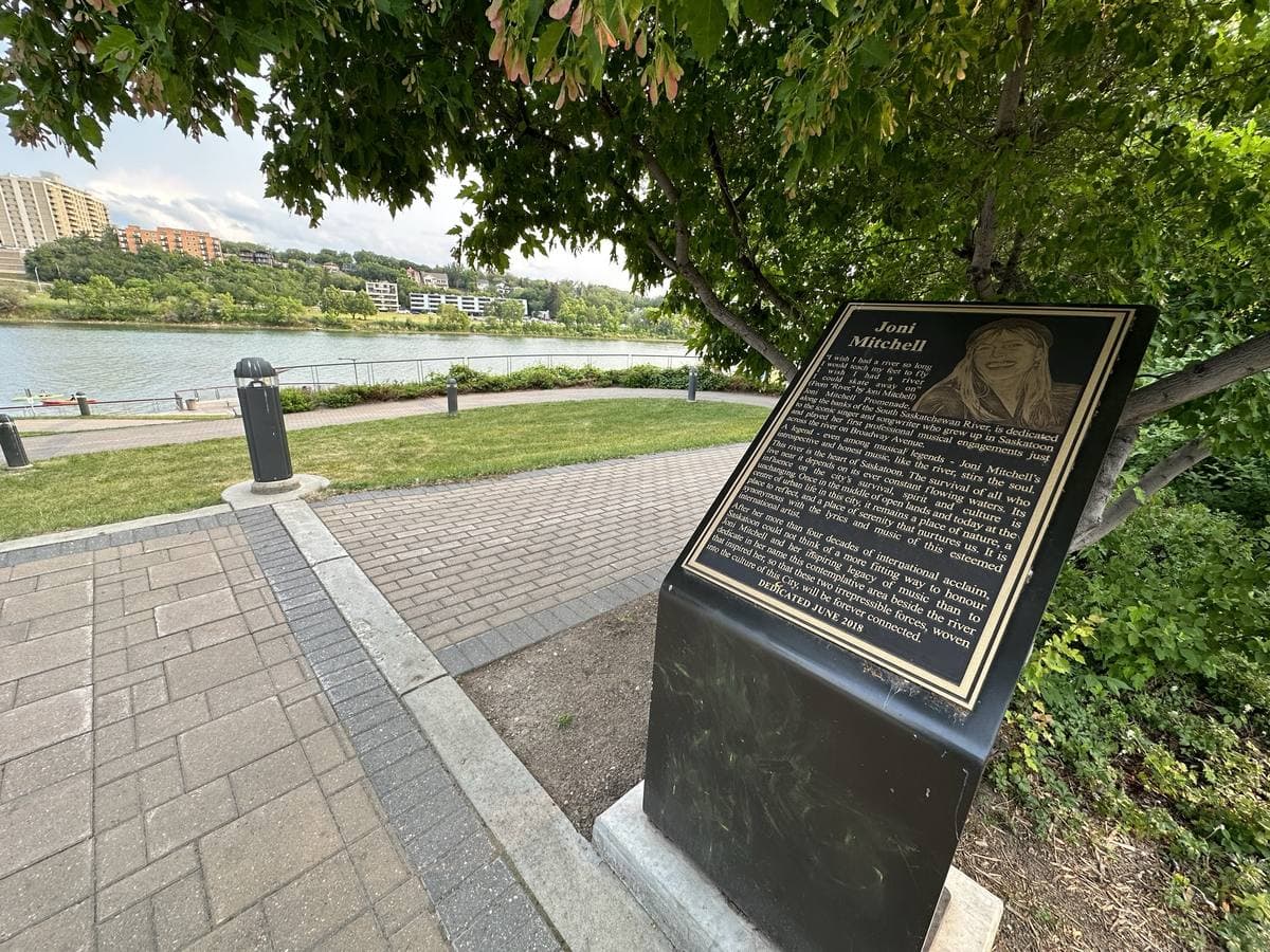 A plaque beside a riverside path, the South Saskatchewan River and a bridge beyond