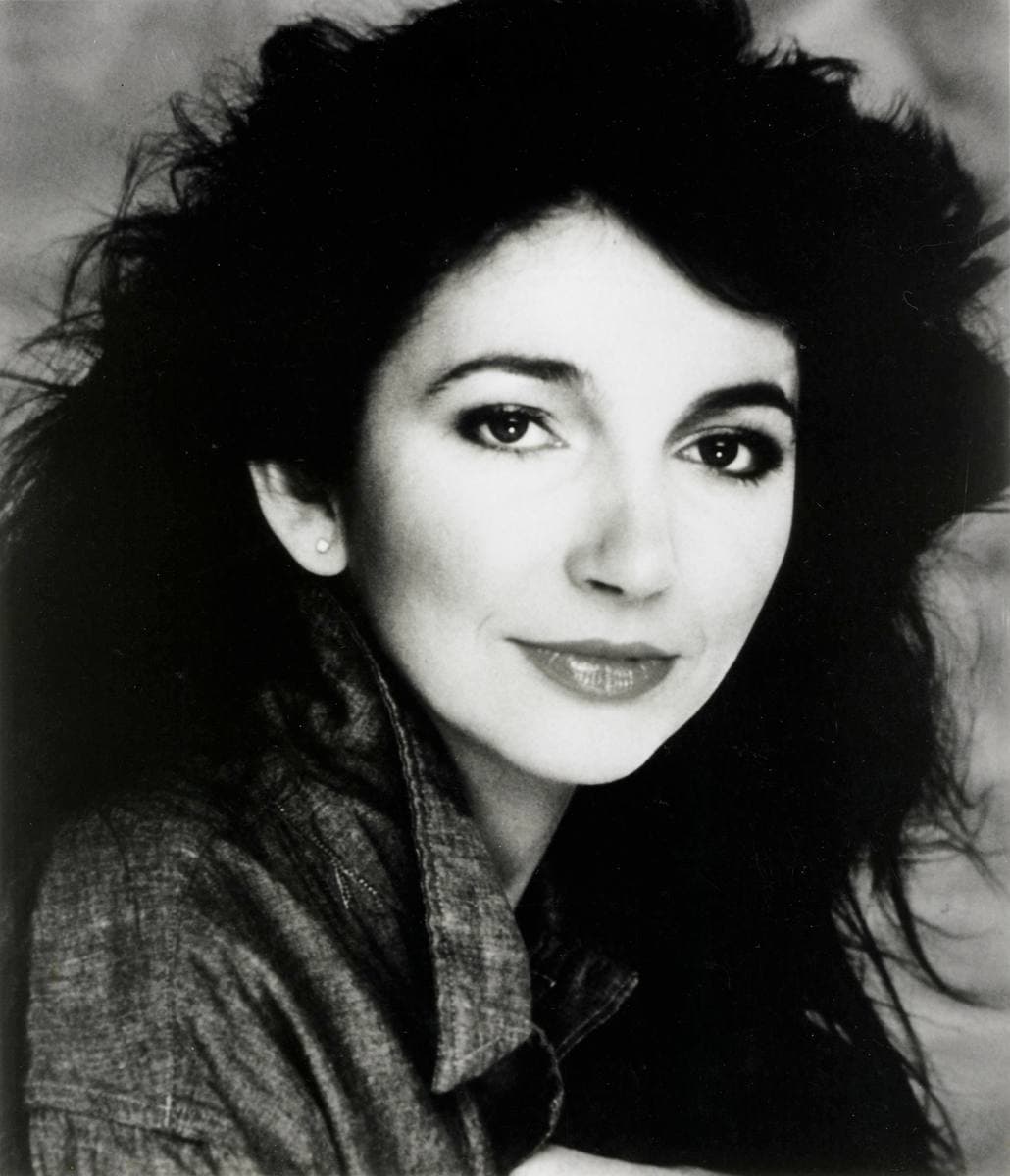 Kate Bush in black and white, head tilted, long dark hair falling around her face, looking straight at the camera with a slight smile