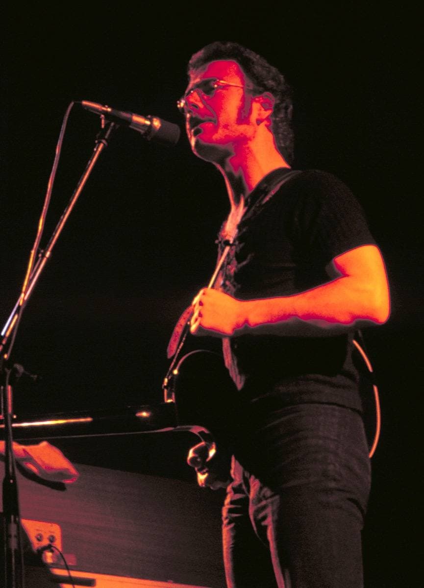 Robert Fripp seated, playing an electric guitar, in glasses