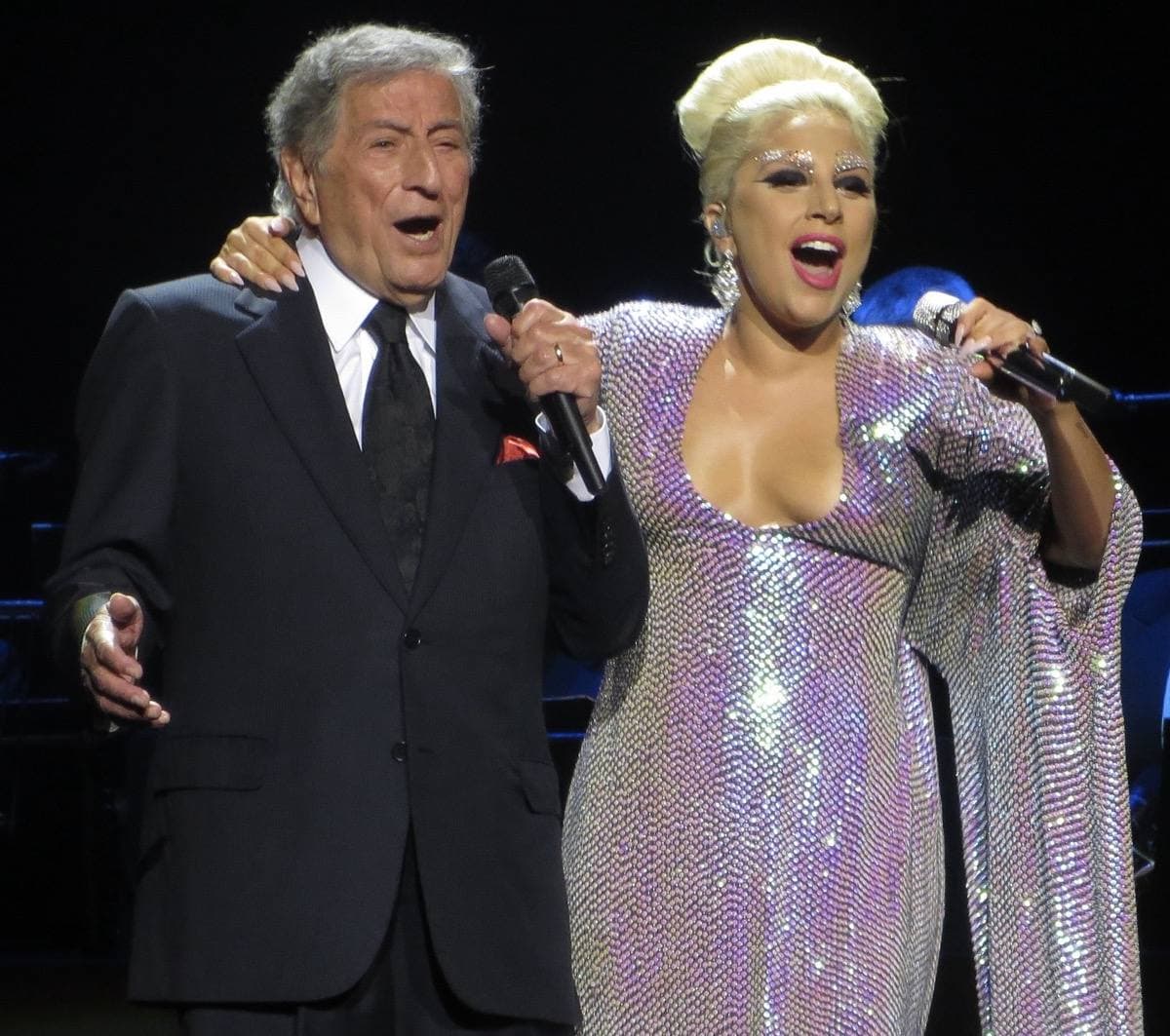 An older man in a dark suit and a woman in a sequinned gown singing together, arm in arm