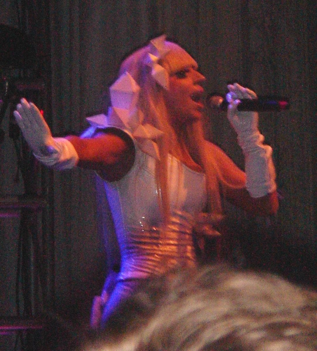 A singer with blonde hair, arm outstretched, singing into a microphone under pink stage light