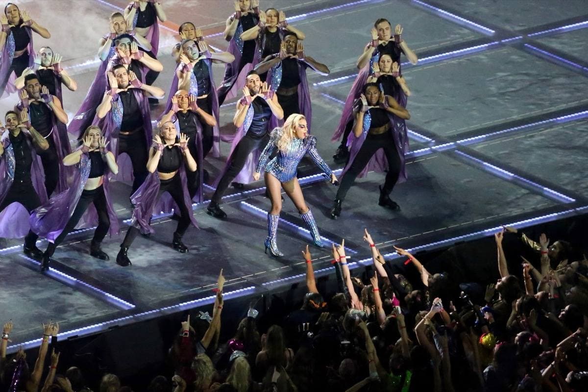 Dancers in formation on a lit stage with the crowd’s raised hands in the foreground