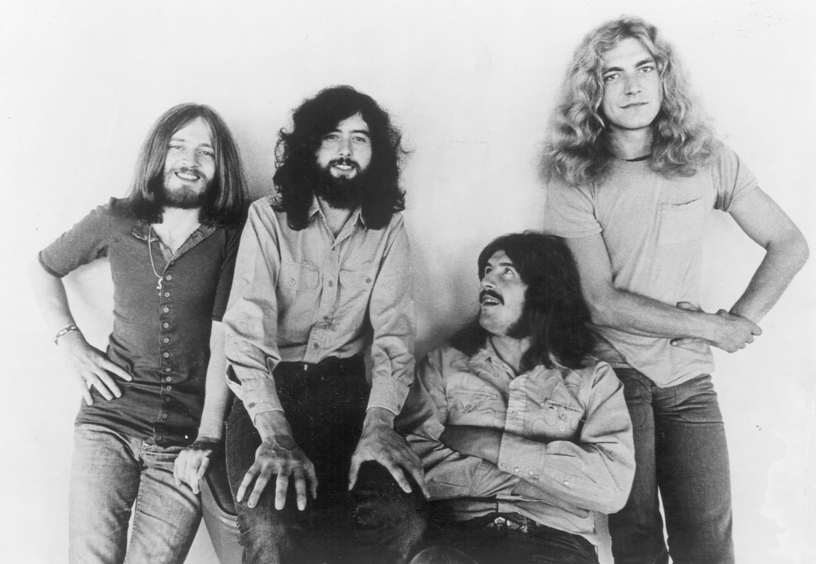 The four members of Led Zeppelin against a pale wall: Jones, Page, Bonham seated, and Plant with his hands on his hips
