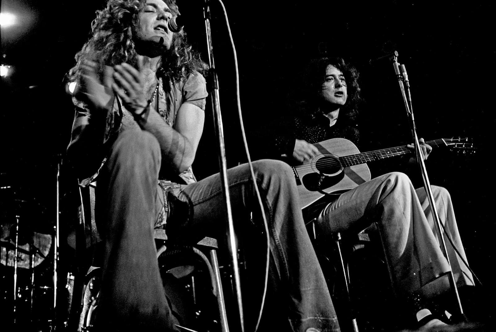 Robert Plant clapping at a microphone beside Jimmy Page seated with an acoustic guitar