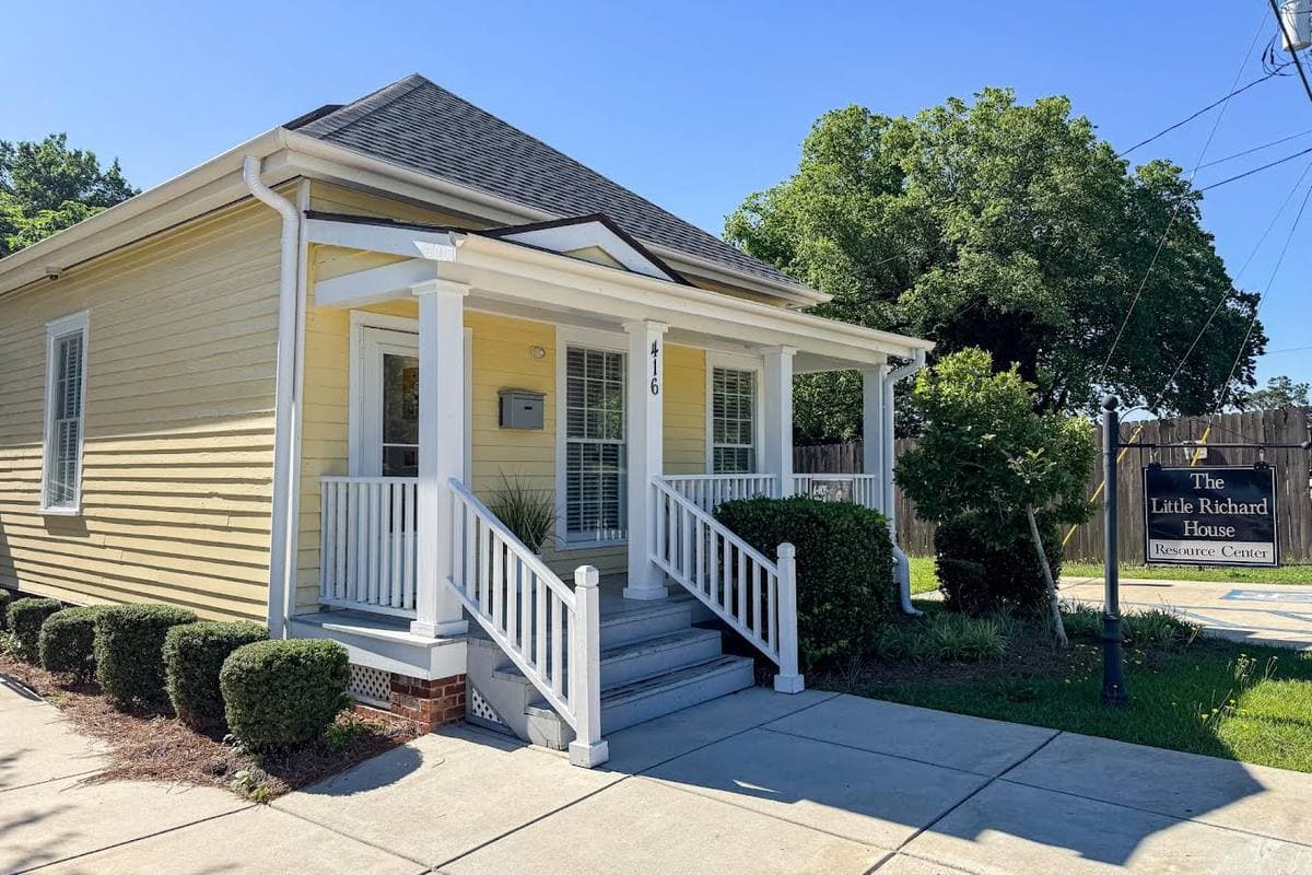 A small pale-yellow wooden house with a white porch and a sign beside it