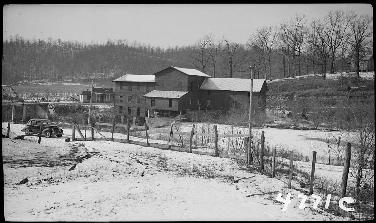 A black-and-white photograph of a wooden mill building by a creek