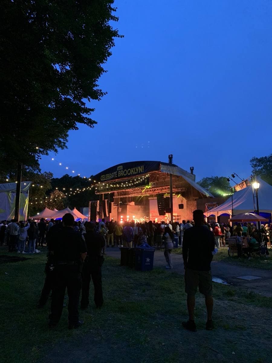 A crowd on a lawn at dusk in front of a lit outdoor stage strung with lights