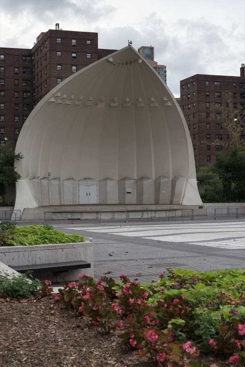 A white shell-shaped bandshell on a paved plaza, with apartment towers behind
