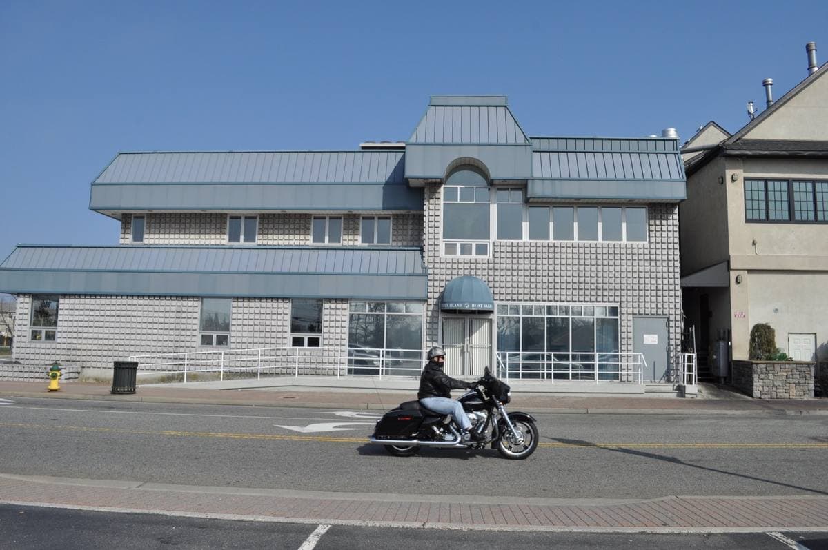 A grey-shingled boat sales building on a waterfront street with a motorcycle passing