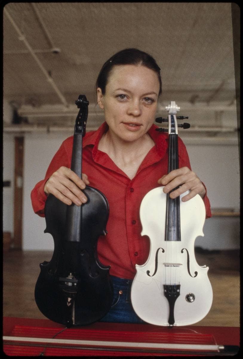 A young woman in a red shirt holding a black violin and a white violin in a bare studio