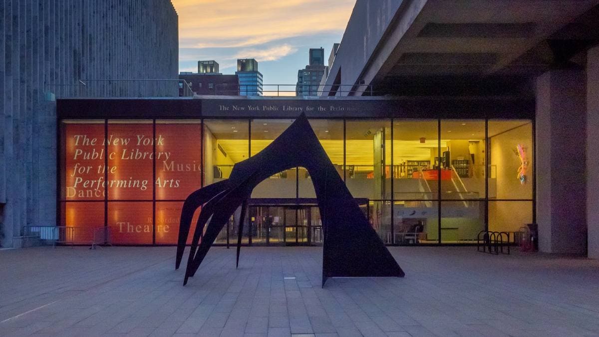 A lit glass library entrance under a concrete overhang, with a black steel sculpture in front at dusk